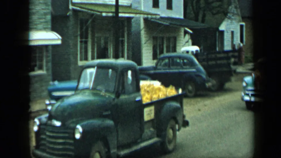 1959 Aledo, Texas street scene with a dark pickup loaded with yellow corn near parked mid-century sedans.