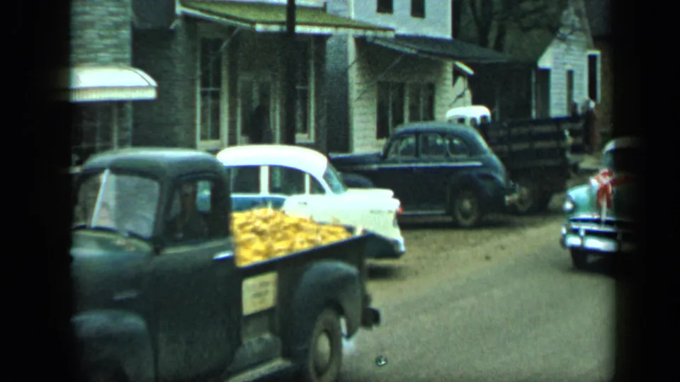 1959 Aledo, Texas frame showing a dark pickup with yellow corn in its bed beside parked cars and storefronts.