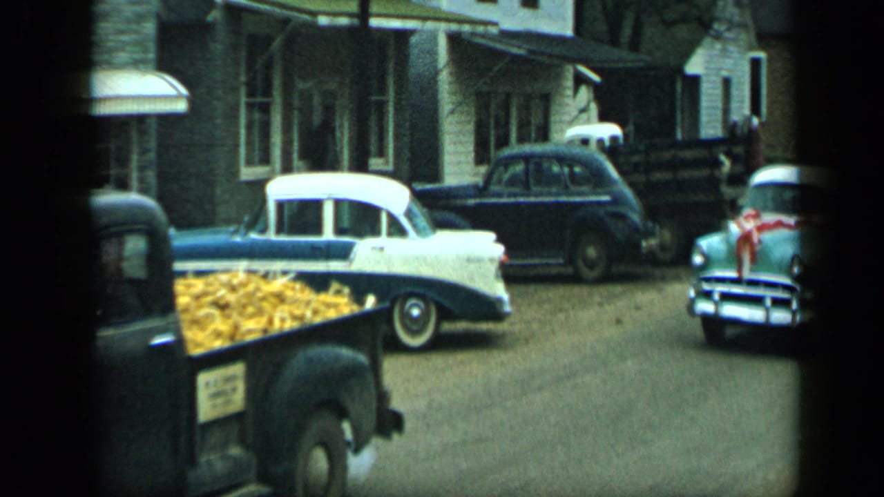 Two-tone sedan parked near a pickup loaded with yellow material on a small town street.