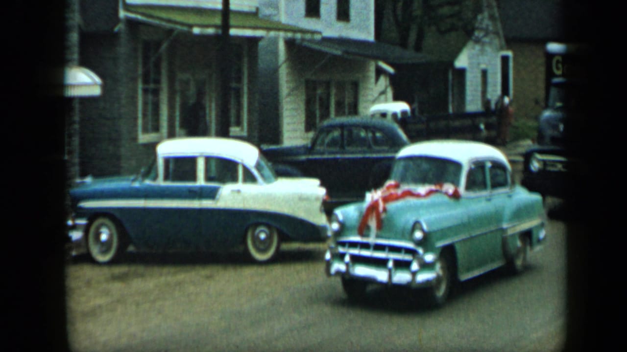 Light green 1950s car with red ribbon passes or turns near a blue-and-white sedan on a street in Aledo, Texas.