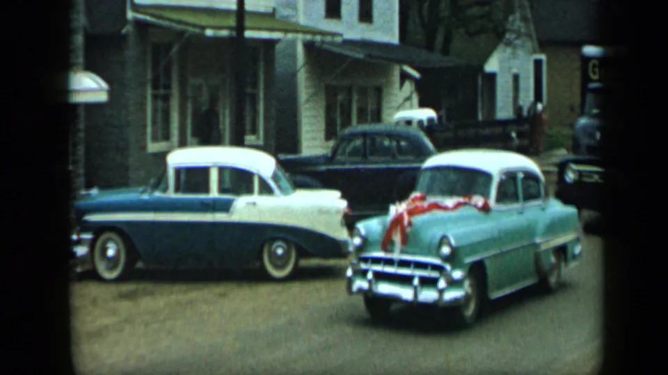Light green 1950s car with red ribbon passes or turns near a blue-and-white sedan on a street in Aledo, Texas.