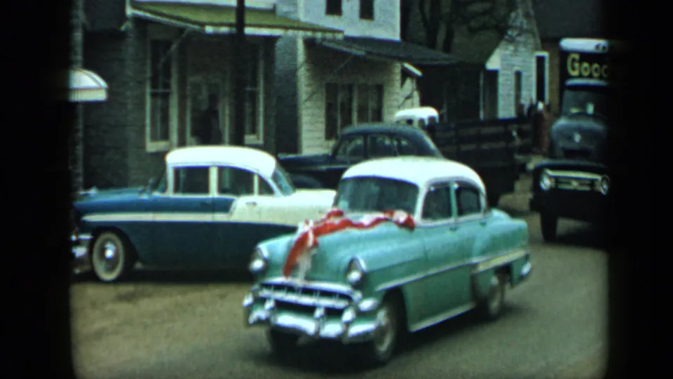 A ribboned light green car fills the foreground while a two-tone sedan, dark sedan, people, and a Goodwill truck appear in the background.