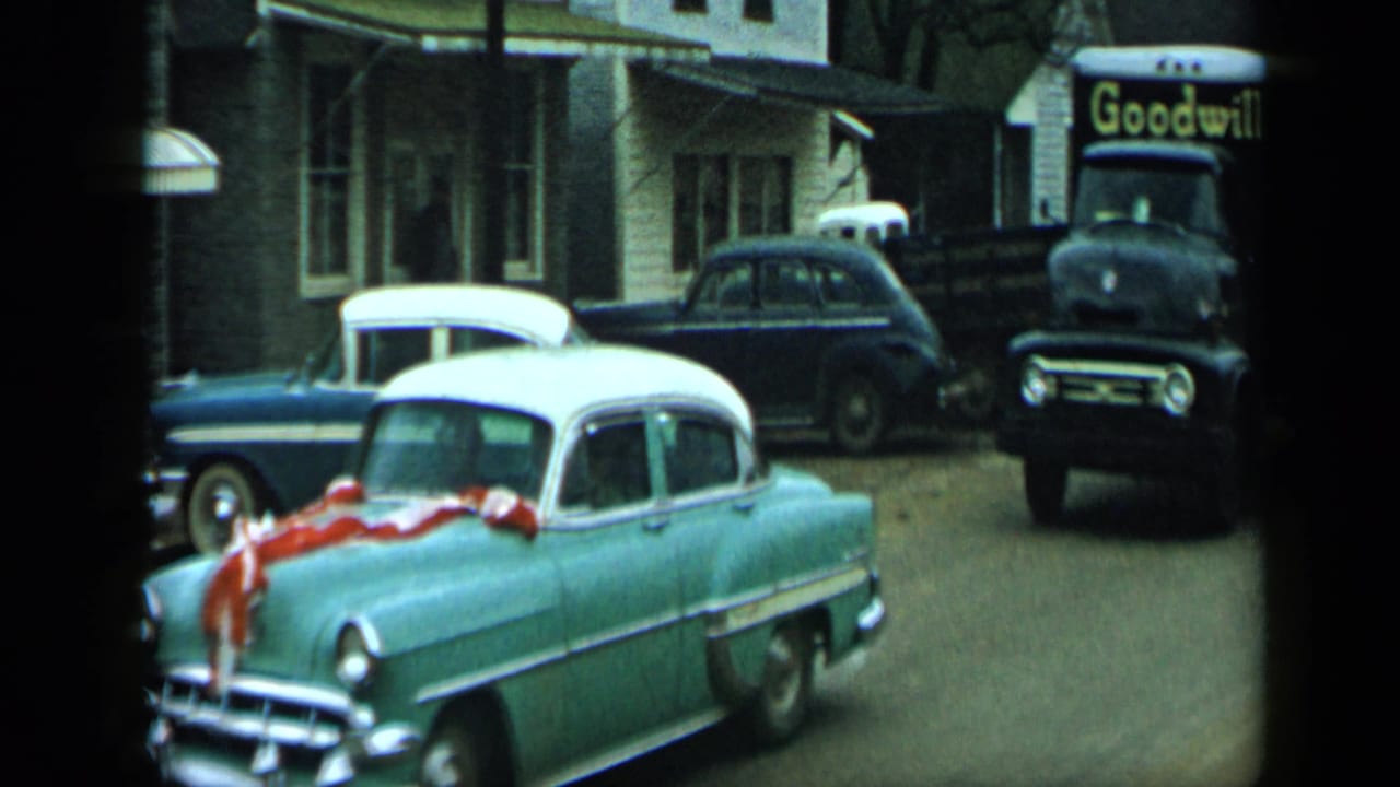 1959 Aledo, Texas street view with a ribboned turquoise-and-white sedan, parked cars, and a Goodwill truck in the background.