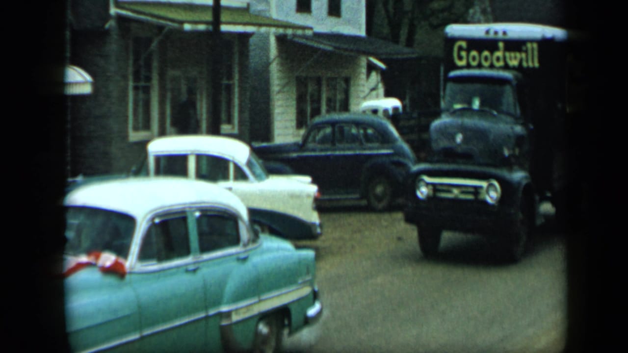 Ribboned turquoise sedan in the foreground with vintage cars and a Goodwill truck behind it.