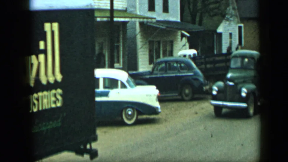 End frame from the 1959 Aledo, Texas footage with the truck partly visible, parked cars, houses, and a distant figure near the sidewalk.