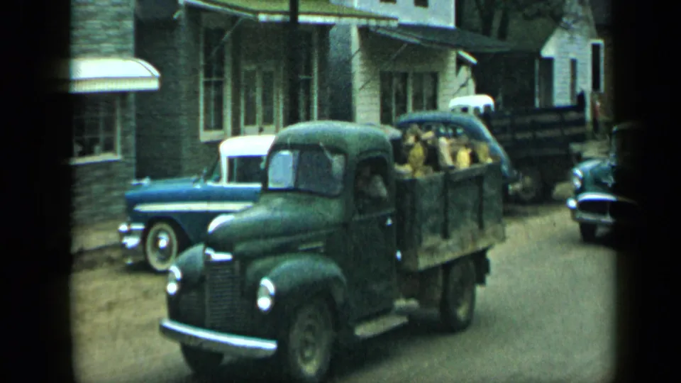 In Aledo, Texas, 1959, a dark green pickup truck moves past parked mid-century cars on a cloudy residential street.