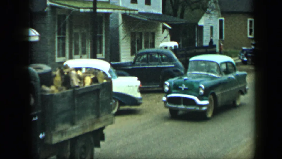 A green mid-century car follows a pickup carrying hay or feed along a quiet residential street in Aledo, Texas, 1959.