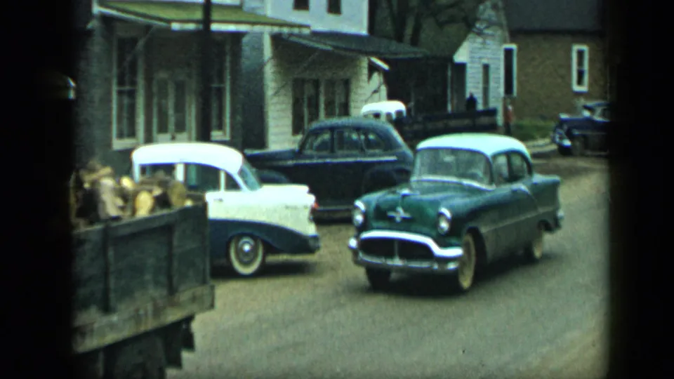 1959 Aledo, Texas home-movie frame showing mid-century cars near houses and a truck bed filled with produce.