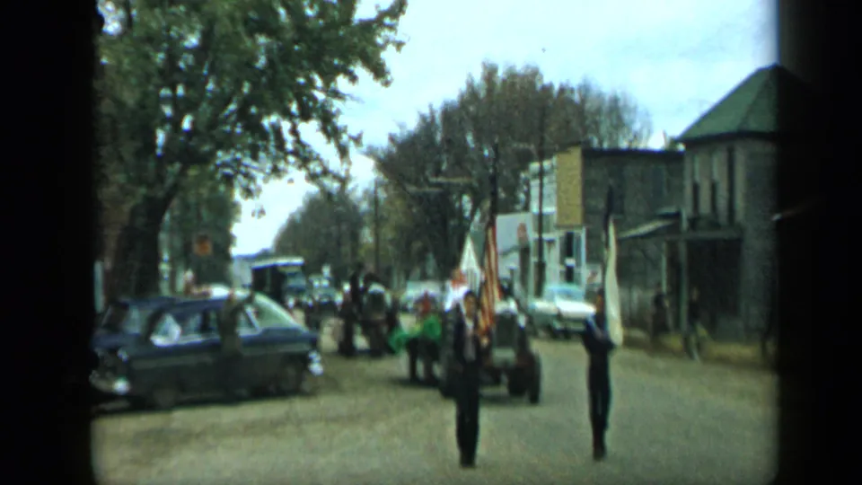 1959 Aledo, Texas home-movie frame showing a small flag procession on a street with vintage cars and storefront-like buildings.