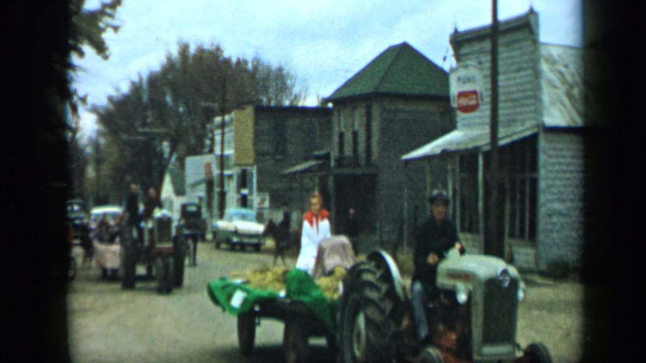 Small-town street with tractors, a hay-filled trailer, a woman in white, storefronts, cars, and pedestrians.