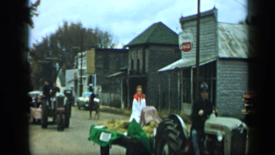Aledo, Texas 1959 home-movie frame with a woman beside a green hay trailer, tractors, a horse, older cars, and storefronts.