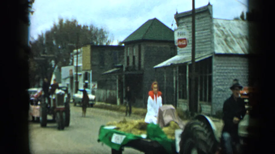 1959 Aledo street view showing a tractor, hay-filled green trailer, woman in white, pedestrians, classic cars, horse, and Coca-Cola sign.
