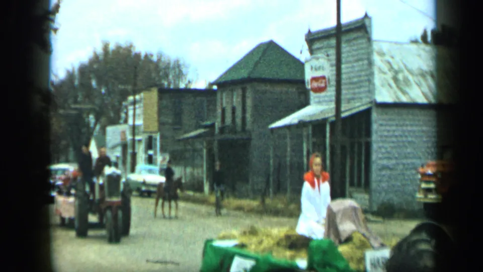 Aledo, Texas 1959 frame of a woman beside a hay trailer as tractors, a horse, pedestrians, cars, and storefronts fill the street.