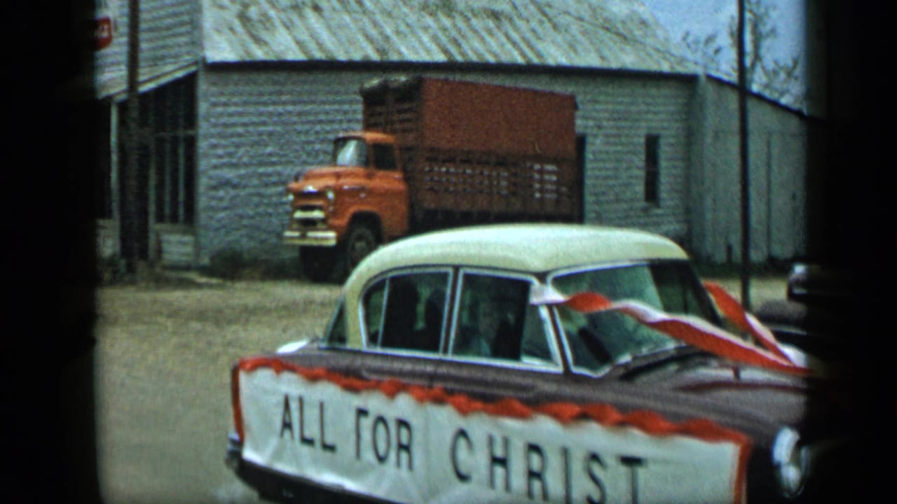 Decorated sedan with ALL FOR CHRIST banner passes in front of a pale building and parked orange truck.