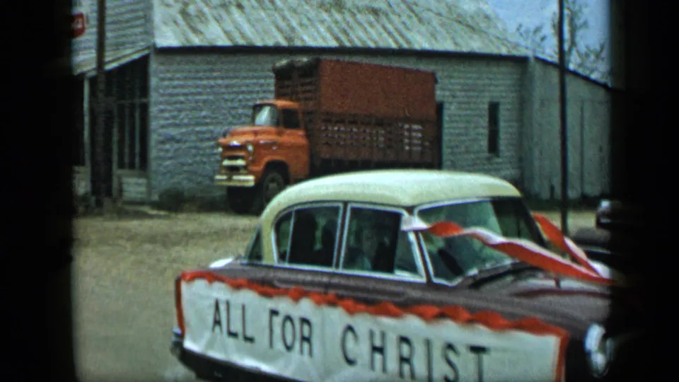 In 1959 Aledo, Texas, a ribboned Chevrolet sedan with an ALL FOR CHRIST banner moves past a pale building and an orange truck.