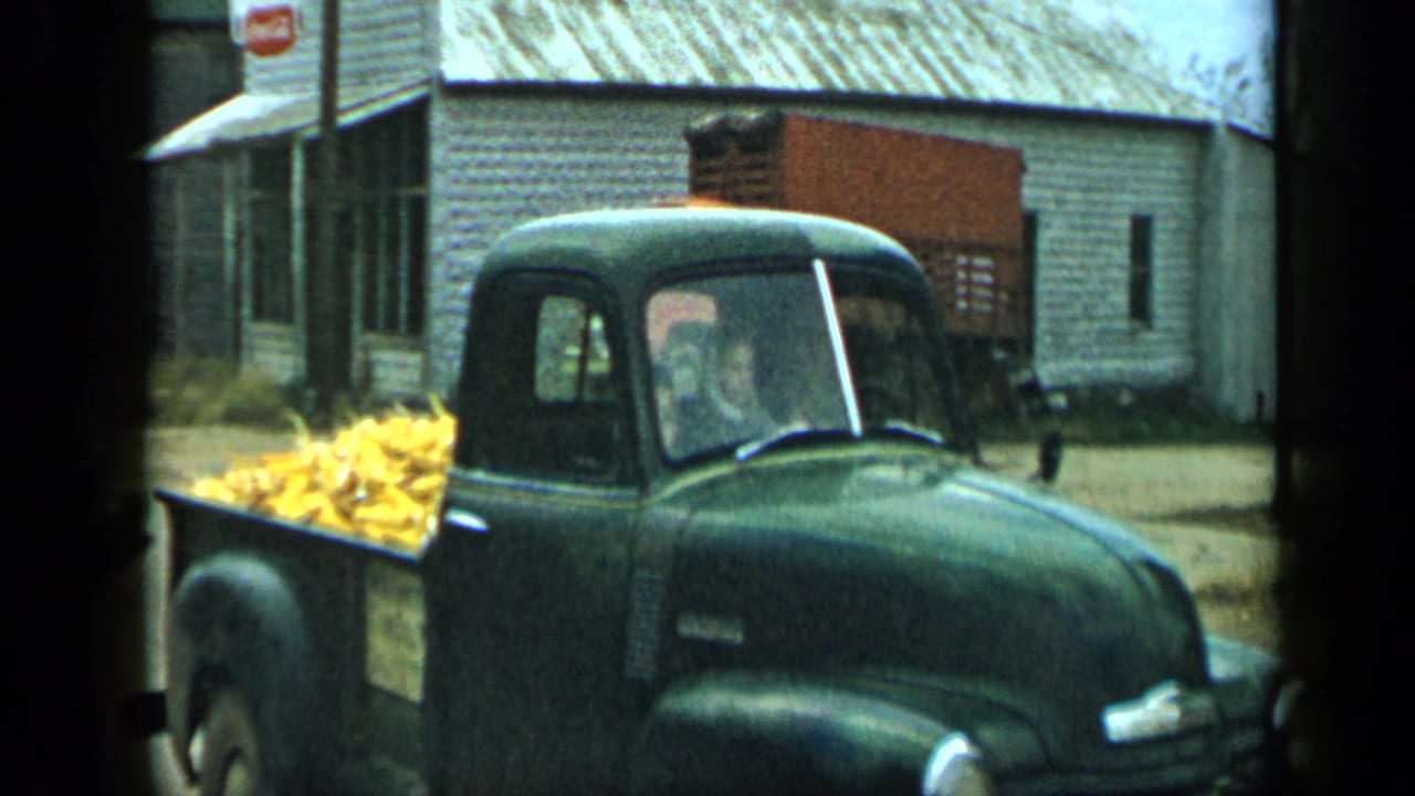 In the Aledo parade footage, a dark green pickup loaded with corn passes close to camera near a pale building and parked orange truck.