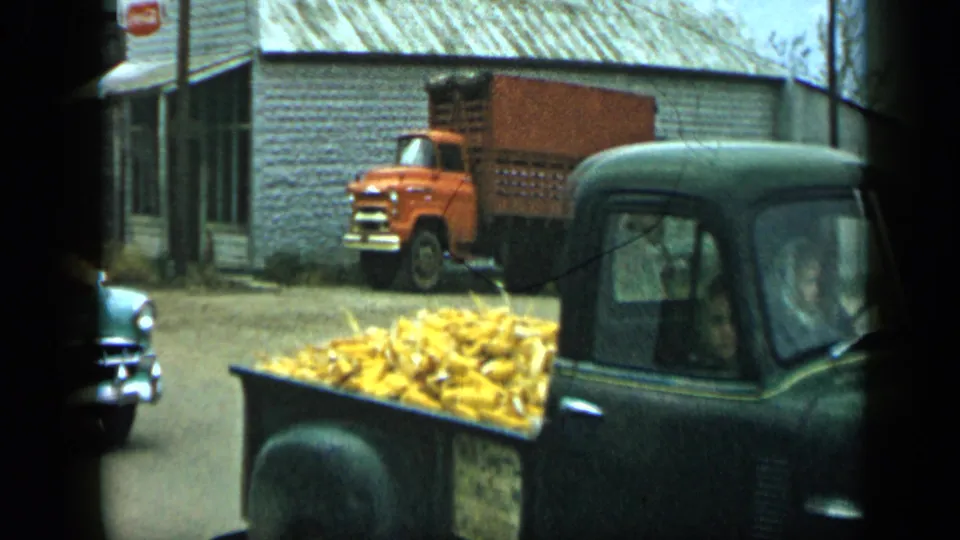 A green pickup with a bed full of corn moves past the camera during the 1959 Aledo, Texas parade, with a parked orange truck behind.