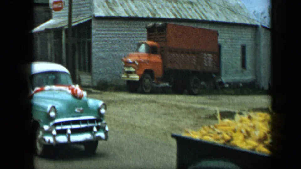 The decorated sedan and corn-loaded pickup continue through the 1959 Aledo, Texas parade area, with an orange truck by a pale building.