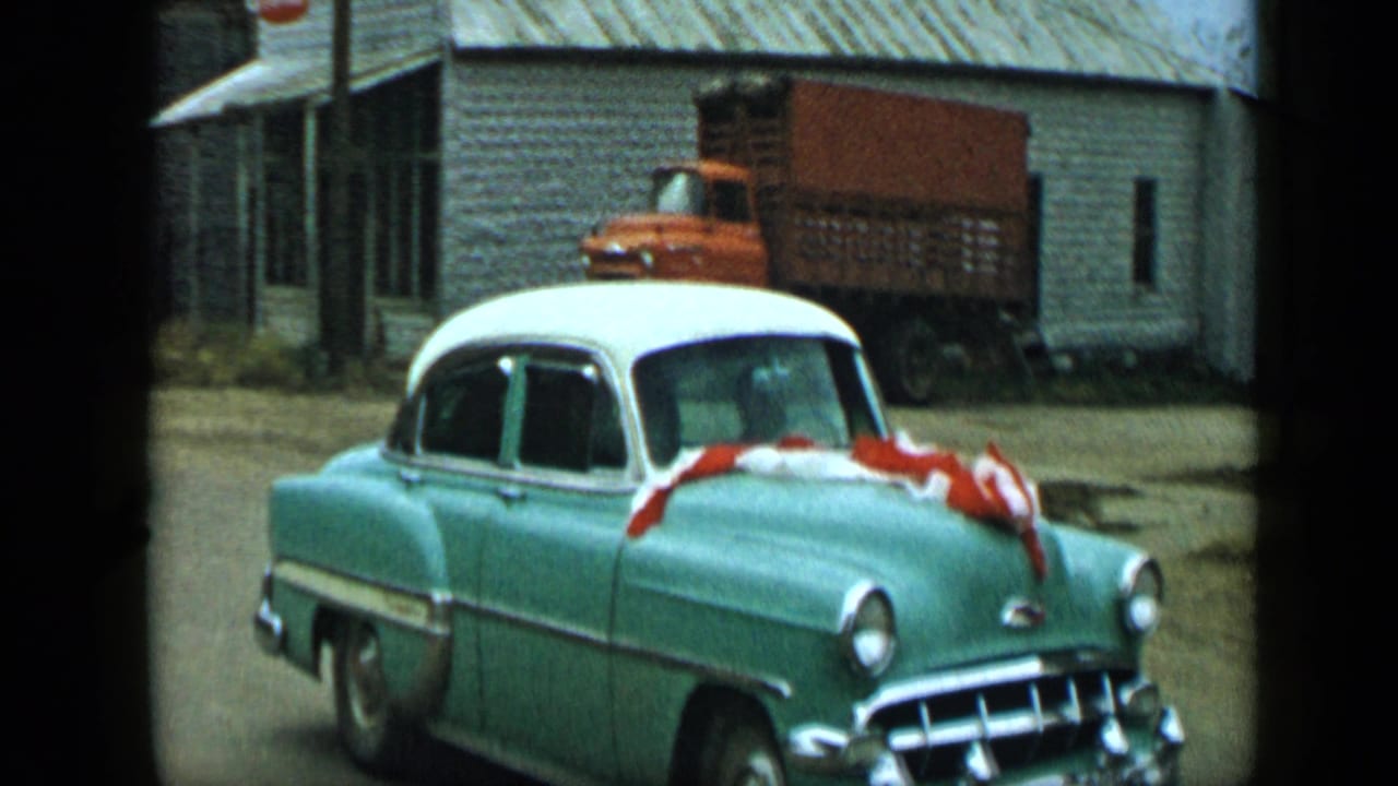 Teal-and-white vintage car with red and white streamers passing a weathered building.