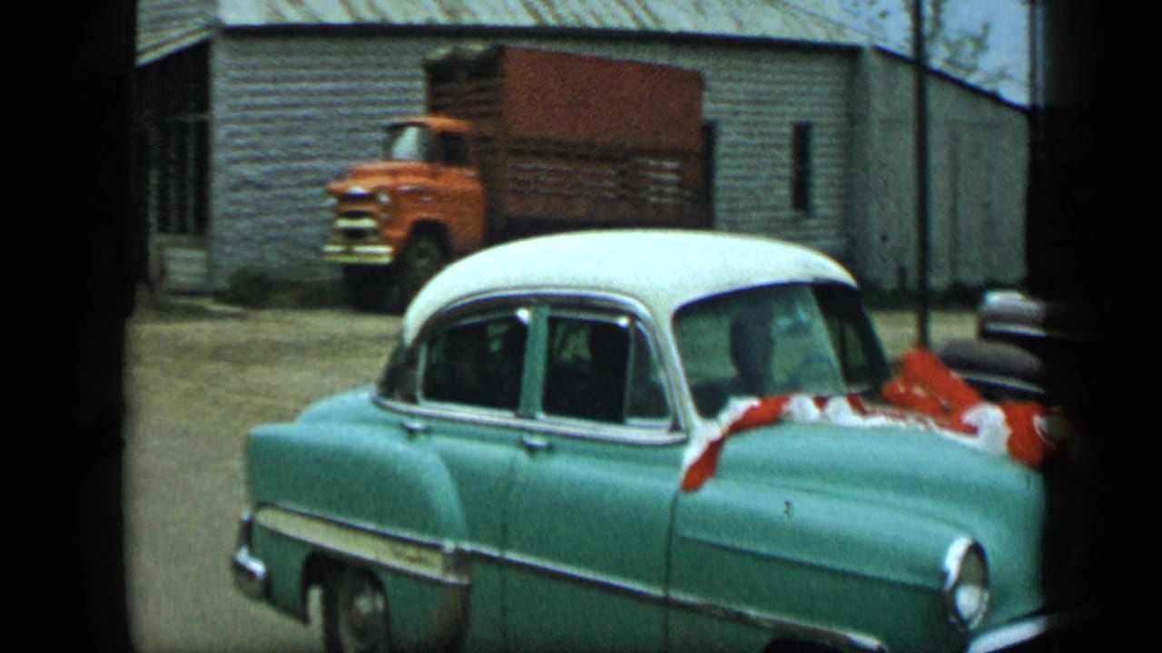 1959 Aledo, Texas frame with a decorated teal-and-white car moving past a building while an orange truck is parked in the background.