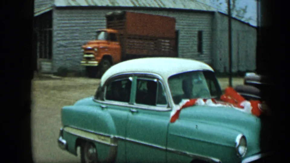 1959 Aledo, Texas frame with a decorated teal-and-white car moving past a building while an orange truck is parked in the background.