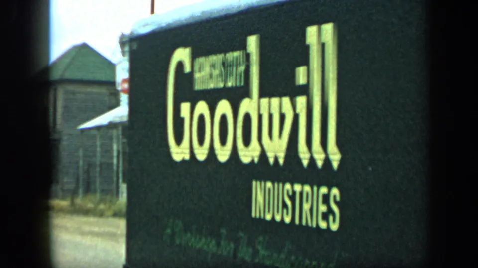 1959 Aledo, Texas frame showing the side of a black Goodwill Industries truck with large yellow lettering near modest roadside buildings.