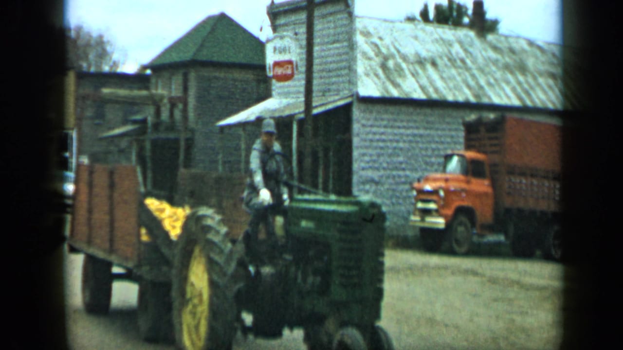 1959 Aledo, Texas frame with a man on a green tractor pulling a wooden trailer of corn past storefronts, a Coca-Cola sign, and an orange truck.