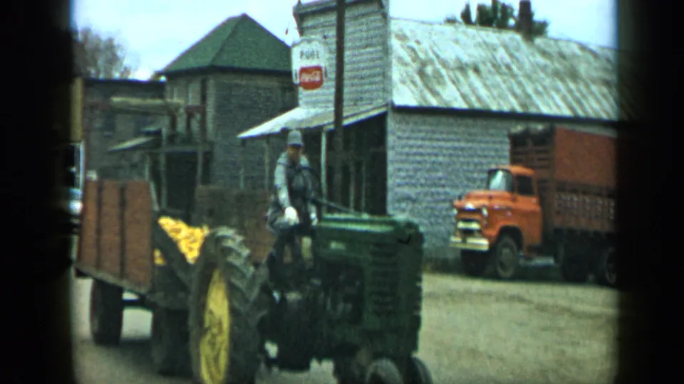 1959 Aledo, Texas frame with a man on a green tractor pulling a wooden trailer of corn past storefronts, a Coca-Cola sign, and an orange truck.