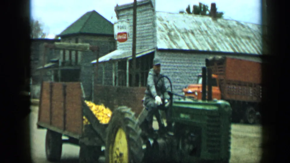 1959 Aledo, Texas frame showing the tractor driver beside a wooden trailer of corn, with storefronts and an orange truck in the background.