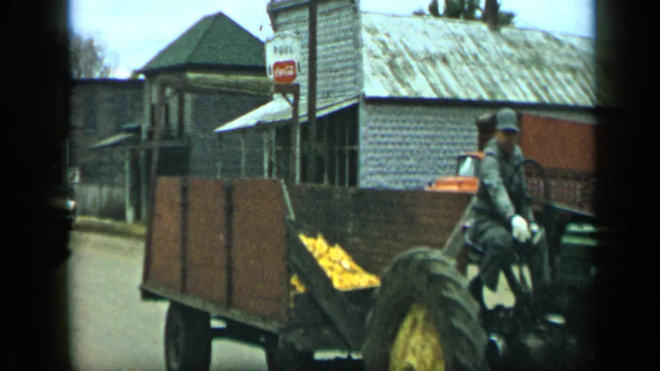 1959 Aledo, Texas frame with a corn-filled wooden trailer and partial tractor driver passing a Coca-Cola sign, storefronts, and an orange truck.