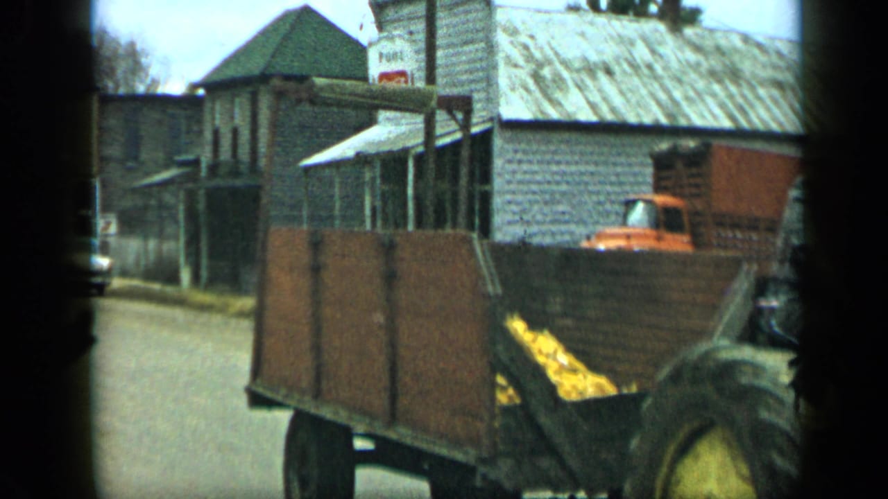 1959 Aledo, Texas home-movie frame showing a tractor and wooden wagon on a street near older buildings and a parked red truck.