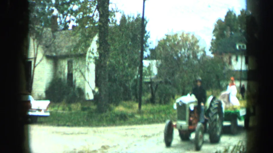 1959 Aledo, Texas frame showing a tractor driver pulling a wagon with riders along a quiet street lined with houses and trees.