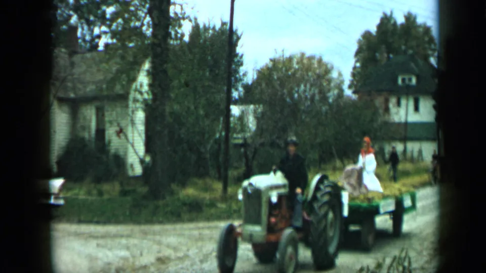 1959 Aledo, Texas home-movie frame of a tractor pulling a wagon with riders, including a person in white and another in red, along a tree-lined street.