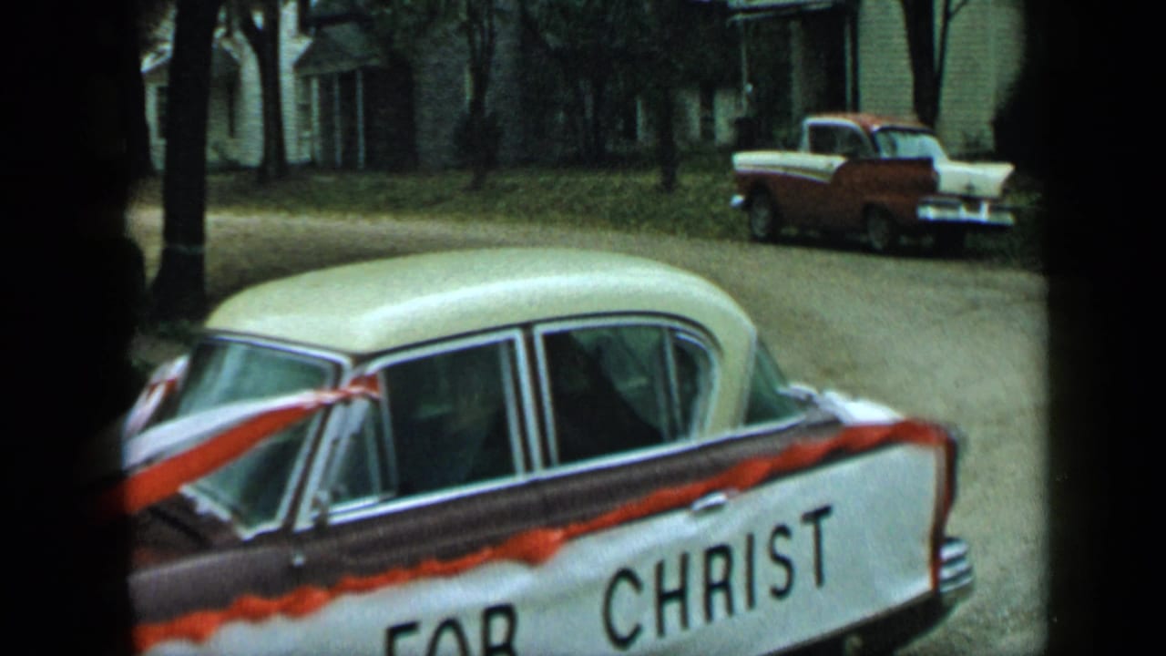 Ribboned sedan marked CHRIST passing a quiet residential street with a pickup in the background.