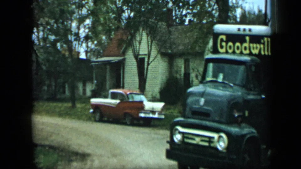 1959 Aledo, Texas frame showing a black Goodwill truck with yellow lettering passing a parked red-and-white pickup near small houses.