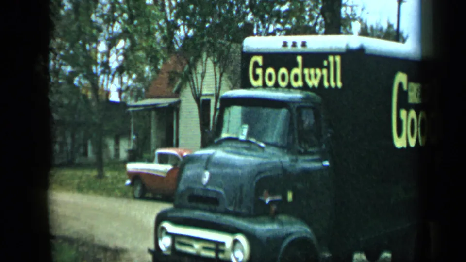 Close 8mm frame of a black Goodwill truck with yellow lettering passing through the Aledo residential street scene in 1959.