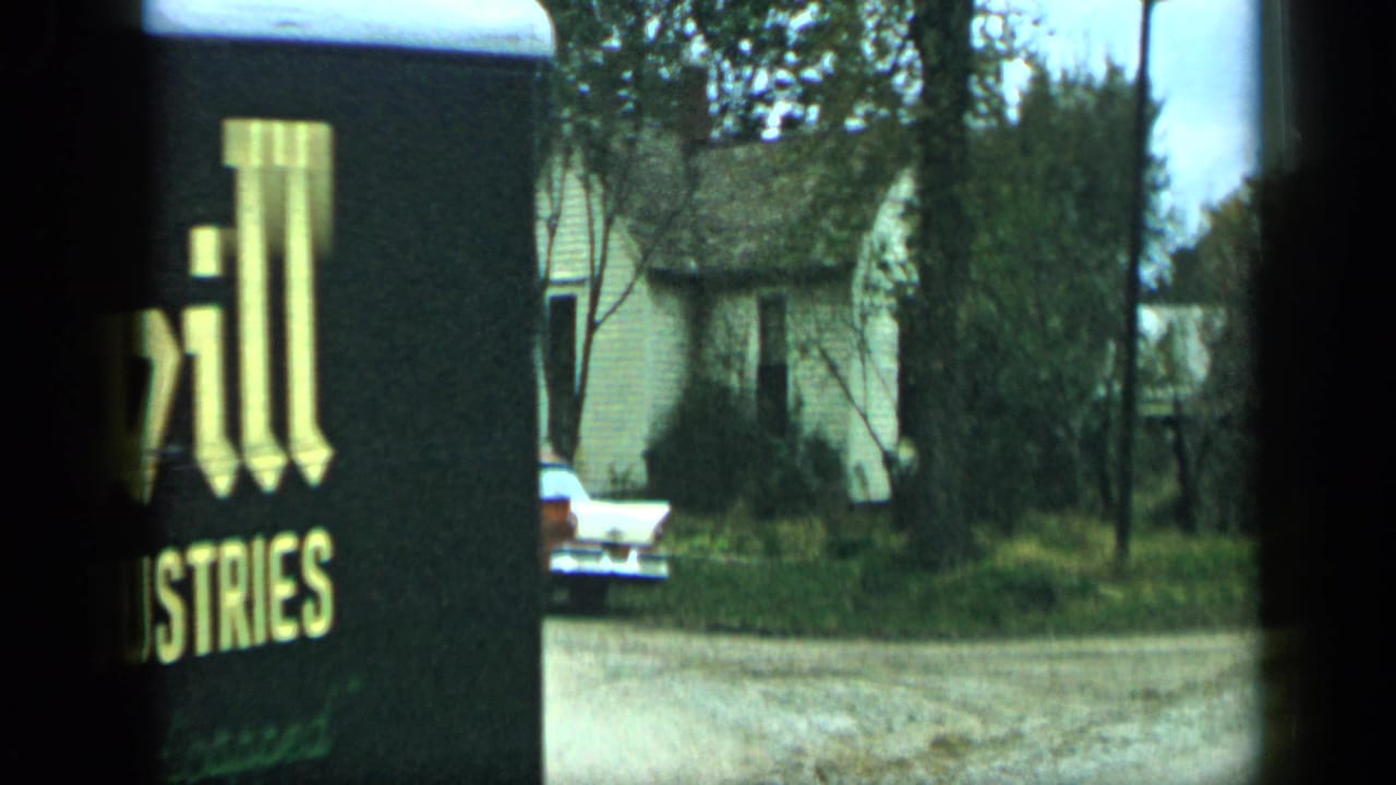 Partial roadside sign beside a residential street with parked cars and houses in the background.