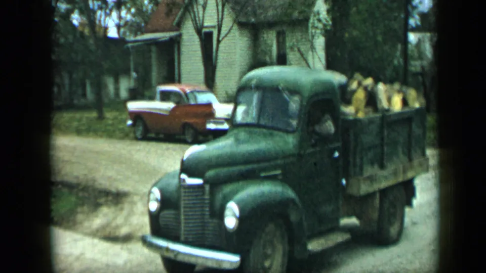 Green flatbed truck carrying firewood passes through a residential street in 1959 Aledo, Texas, with a red-and-white sedan nearby.