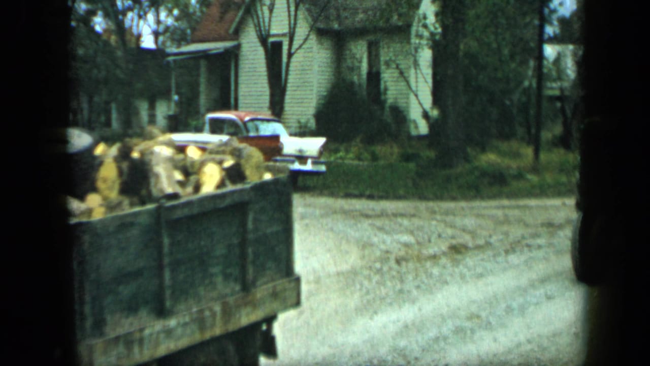 Tractor wagon loaded with corn near a gravel road, with a two-tone car and house in the background.