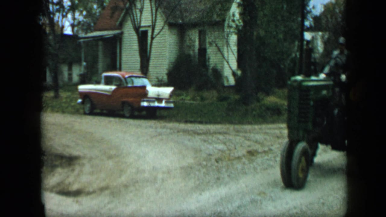 Red-and-white sedan parked near a house as a green tractor enters from the right edge.