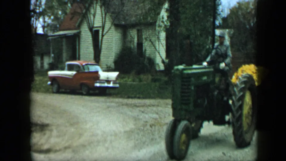 1959 Aledo, Texas frame showing a man operating a green tractor beside a gravel road and parked two-tone sedan.