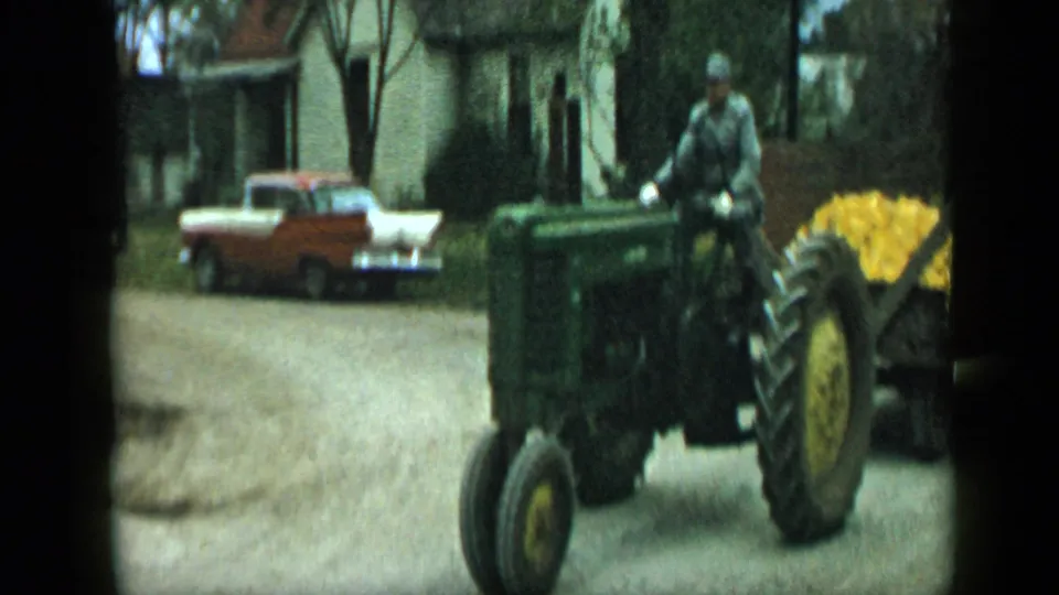 1959 Aledo, Texas frame of a green tractor pulling a yellow corn load past a parked sedan and roadside house.