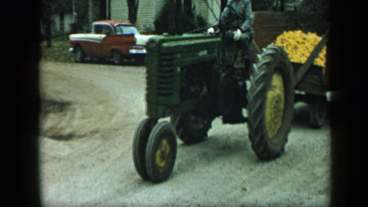 Green tractor and corn trailer pass on a dirt road near a parked two-tone car.