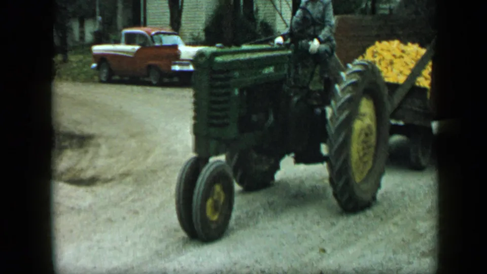 In Aledo, Texas, 1959, a green tractor pulling a wooden trailer of yellow corn moves past a red-and-white parked car on a dirt road.