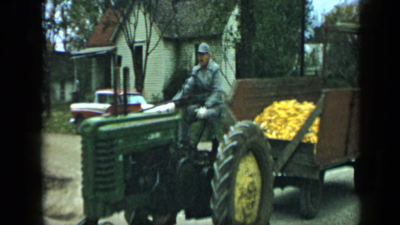 Man in a light uniform drives a green tractor pulling a wooden corn trailer.