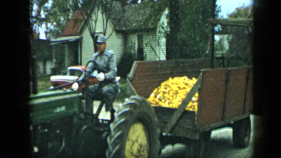 The uniformed driver passes close to the handheld camera on a green tractor, with a wooden corn trailer and rural houses behind him.