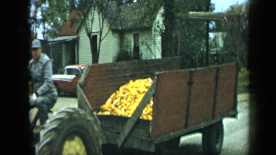 The corn-loaded wooden trailer trails the tractor through the 1959 Aledo street scene, with a white house and two-tone car in the background.