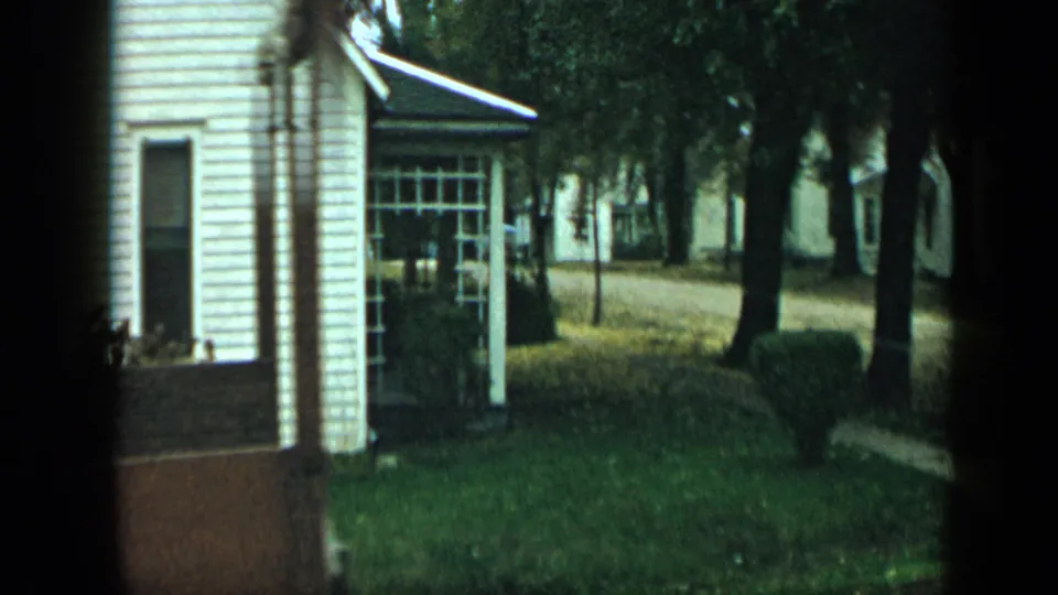 Residential Aledo street view with a white clapboard house, porch, lawn, trees, and the edge of a wooden trailer in 8mm footage.
