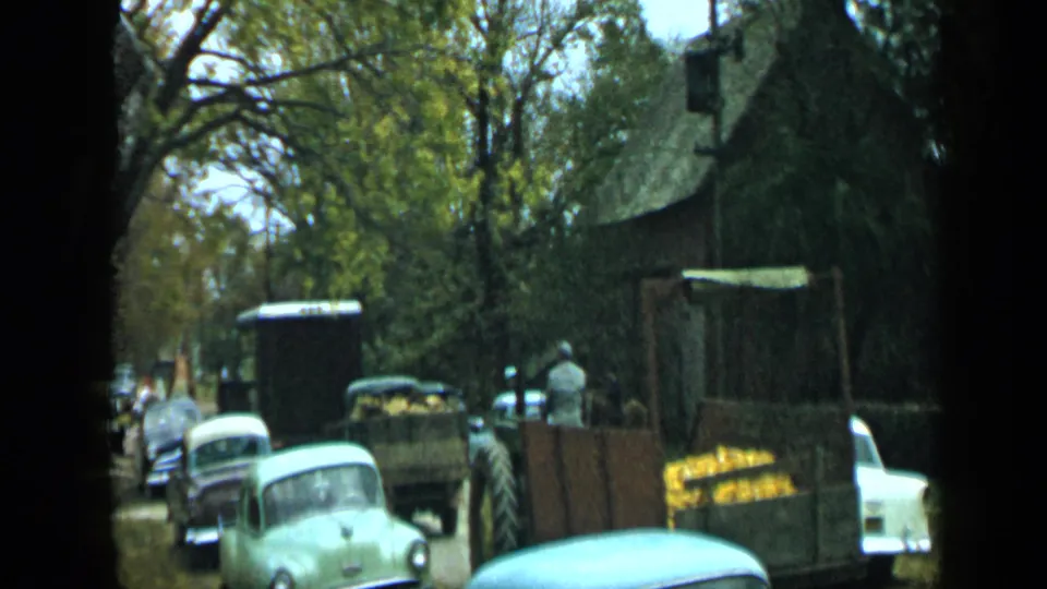 1959 Aledo, Texas street scene with a tractor pulling or standing by a wooden produce trailer near classic cars and trees.