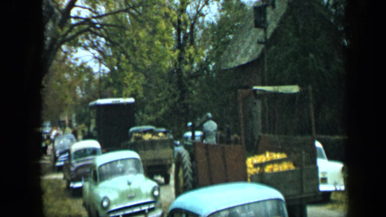 Tree-lined street with parked vintage cars and a farm trailer carrying yellow produce.